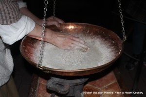 After the twelfth charge of sugar on the caraway seeds for the receipt (recipe) Carraway Comfits, the sugar coated seeds are moved around the hot, swinging comfit pan by hand until all the caraway seeds feel dry.  Now the caraway seeds have twelve coats of sugar and must rest for another day of charges until the total coats is sixty.