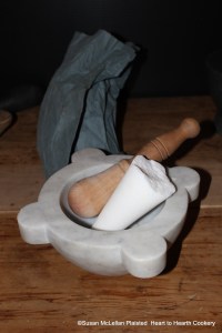 After the cacao nuts (raw fermented cacao beans) have been finely beaten and searced for the receipt (recipe) To make Chocolate do the same with half a pound of hard sugar.  Pictured is the top I knocked off from a sugar loaf in my marble mortar and pestle that weighed a quarter pound as I was making half the receipt.  A loaf of sugar is wrapped in blue paper behind the marble mortar.