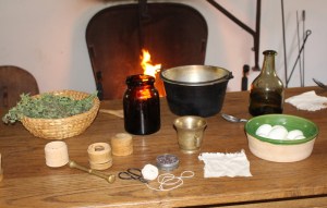 All ingredients and equipment are ready on the table for the preparation of the 1777 receipt (recipe) To Pickle Eggs.  Spices, cochineal, brass mortar and pestle, rag, hard boiled eggs in the front and basket of sweet herbs, glass preserving jar, preserving kettle, and white wine vinegar in the back