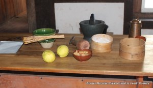 Pictured is the prep table for the receipt (recipe) Quince Puffs: My Lady Sheldons Receipt. From left to right, the baking tin lined with white paper, birch twig whisk and bowl for preparing the egg white. two quince, small bowl with the boiled quince, wood masher, marble mortar and pestle, sugar measure and sieve.