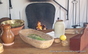 The table is ready with  the ingredients and equipment needed to prepare the receipt (recipe) To preserve Asparagus - red ware pitcher of water, colander, rye straw basket of asparagus, cloves, lemon, salt box and salt-glazed pot sitting on the napkin.